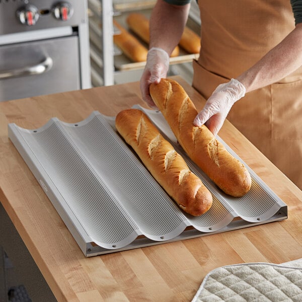 A person holding two long loaves of bread on a Baker's Lane baguette pan.