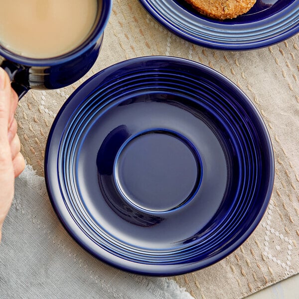 A hand holding a cup of coffee and a blue Acopa Capri saucer on a table.