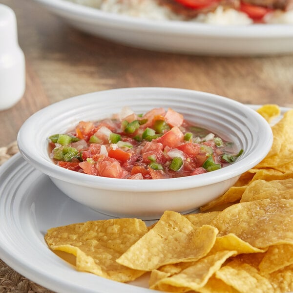 A white stoneware bowl of salsa on a table with a plate of chips.