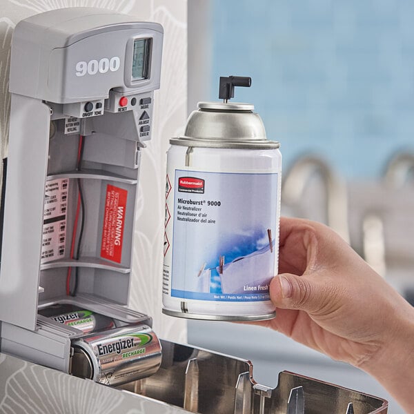 A hand holding a Rubbermaid Microburst 9000 Linen Fresh air freshener spray can near a dispenser.