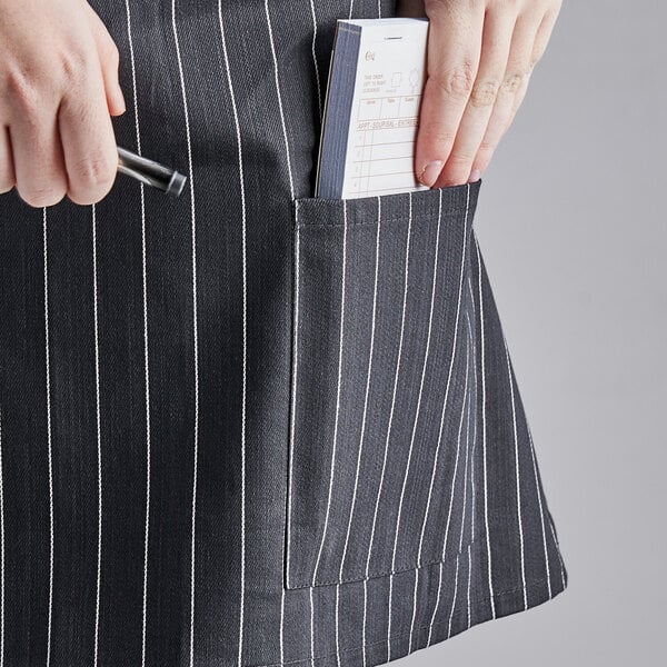 A person holding a pen in a pocket of a black and white pinstripe half bistro apron.