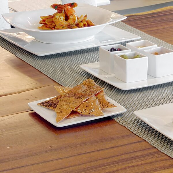 A table with a variety of food on white square Front of the House porcelain plates.