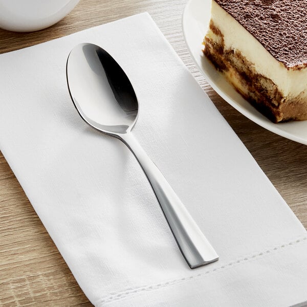 An Acopa stainless steel spoon on a white napkin next to a plate of dessert.