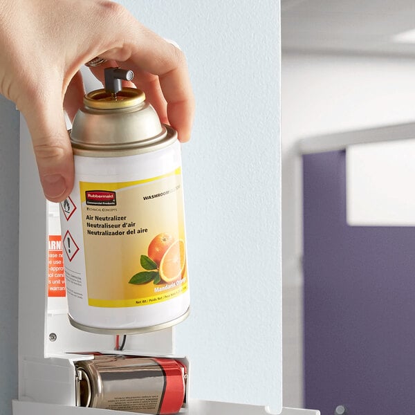 A hand using a Rubbermaid Mandarin Orange air freshener refill to spray air freshener in a room.