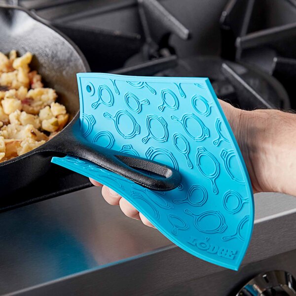 A hand holding a turquoise Lodge silicone trivet over a pan with a blue silicone spatula.