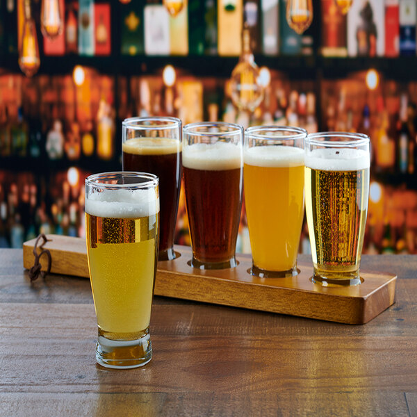 A wooden flight paddle holding five glasses of beer in various colors, with one glass placed in front on a wooden surface.