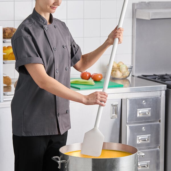 A long white paddle with a polyethylene blade and polypropylene handle being used to stir a large pot in a commercial kitchen.