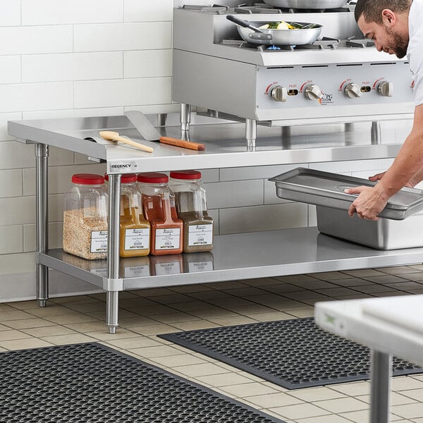 A man in a professional kitchen using a Regency stainless steel equipment stand with an undershelf to hold a metal container.