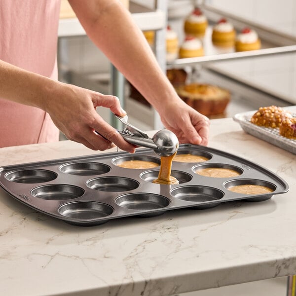A non-stick 12 mold muffin top or slider bun pan being filled with batter using a scoop.