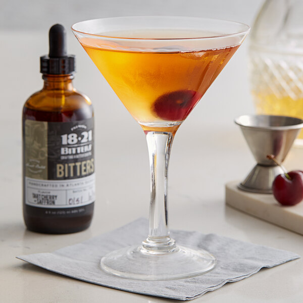 A glass of a cocktail with a cherry and a bottle of 18.21 Tart Cherry & Saffron Bitters on a table.