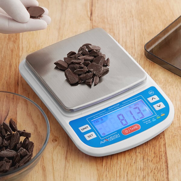 A hand using an Avaweigh digital portion scale to weigh a pile of chocolate pieces.