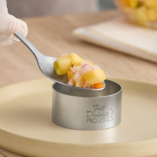 A stainless steel round cake and food ring mold labeled 'Fat Daddio's ProSeries' being used to shape food on a plate.