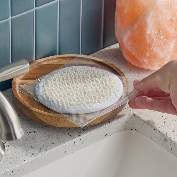 A round, white bath loofah in a clear plastic wrapper placed on a wooden dish near a bathroom sink.