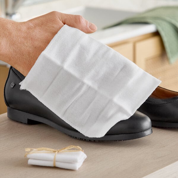 A white shoe mitt being used to clean a black shoe, with a stack of similar mitts tied with a natural ribbon nearby.