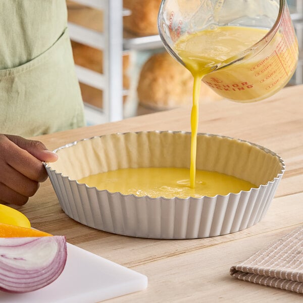 A round, deep fluted tart or quiche pan with a removable bottom being filled with a yellow liquid mixture.