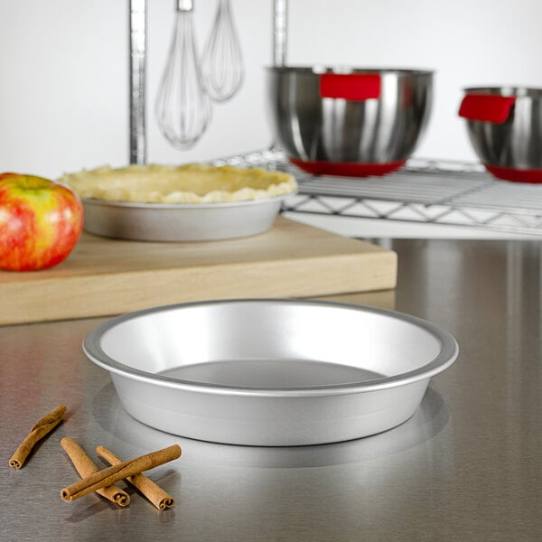 A silver Fat Daddio's ProSeries pie pan on a counter with red and yellow apples and cinnamon sticks.