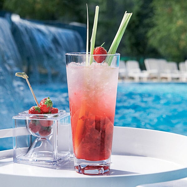 A clear Arcoroc SAN plastic hi ball glass with a red drink and strawberries on a table by a pool.