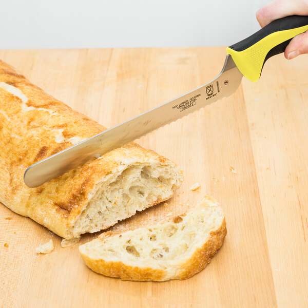 A hand holding a yellow Mercer Culinary bread knife cutting a loaf of bread.