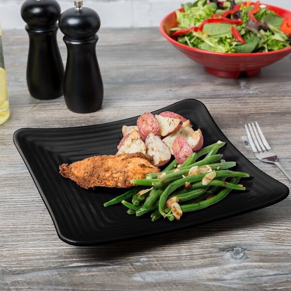 A black Milano melamine square plate with chicken and green beans on a table.