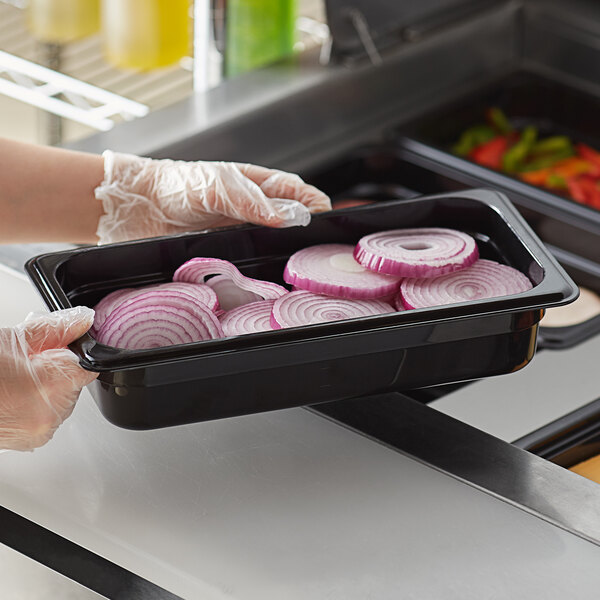 A gloved hand using a Vigor black polycarbonate food pan to hold onions.