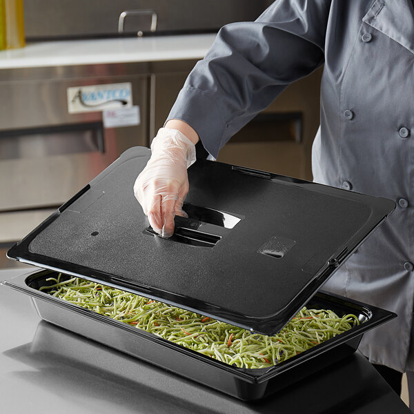 A person with a chef's uniform using a Vigor black polycarbonate food pan lid to cover a tray of food.