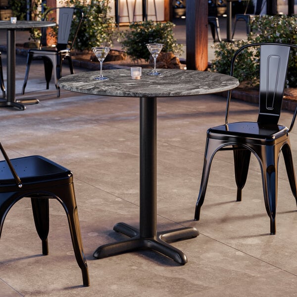 A round table with a smooth Paladina finish and a black pedestal base, set outdoors with two black metal chairs.