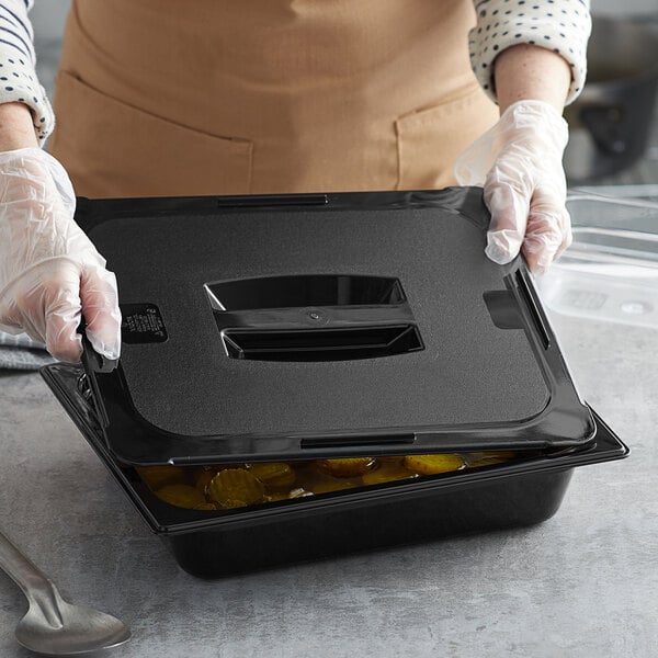 A woman in gloves using a Vigor black polycarbonate food pan lid to cover a black container of food.