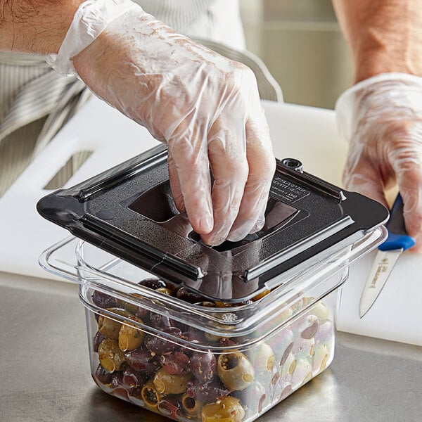 A person wearing a plastic glove in a close-up opening a Vigor black plastic food container with olives.