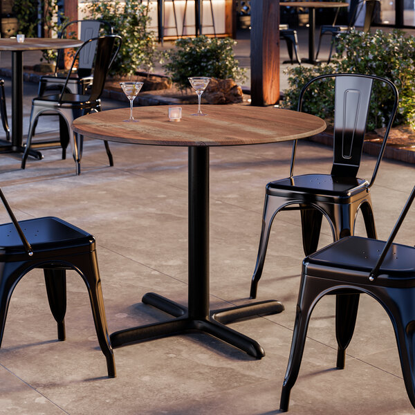 A round table with a textured Yukon oak finish and a black pedestal base, surrounded by black metal chairs.