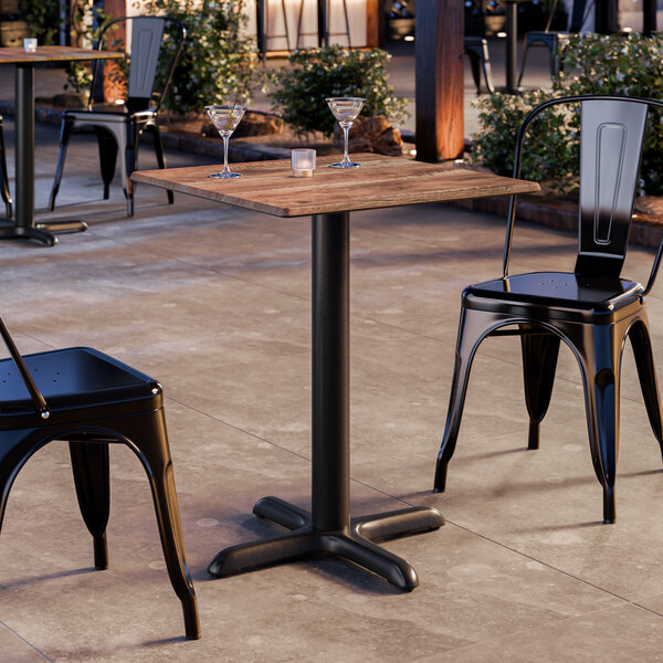 A square table with a textured Yukon oak finish and a black pedestal base, set outdoors with two black metal chairs.