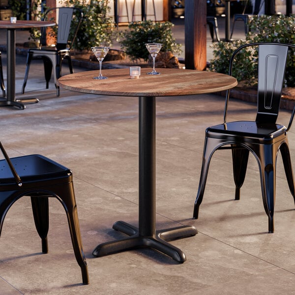 A round table with a textured Yukon oak finish and a black pedestal base, set outdoors with two black metal chairs.