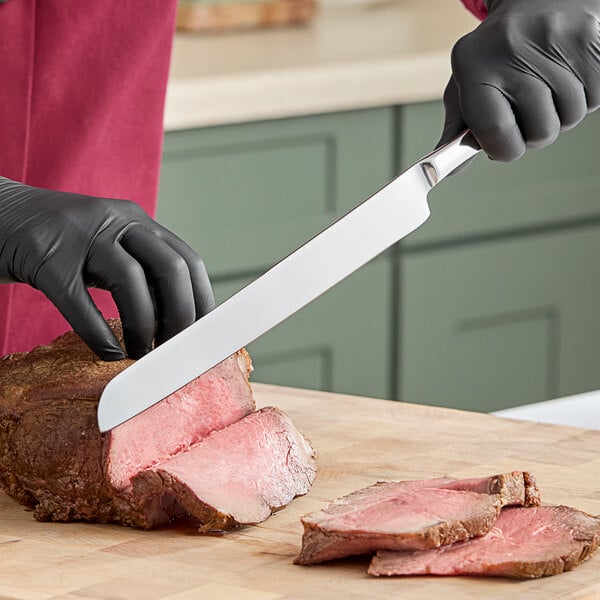 A gloved hand uses a Fortessa Grand City stainless steel carving knife to cut meat on a wooden cutting board.