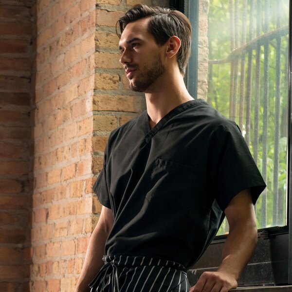 A man wearing a black Uncommon Chef cook shirt leaning against a window.