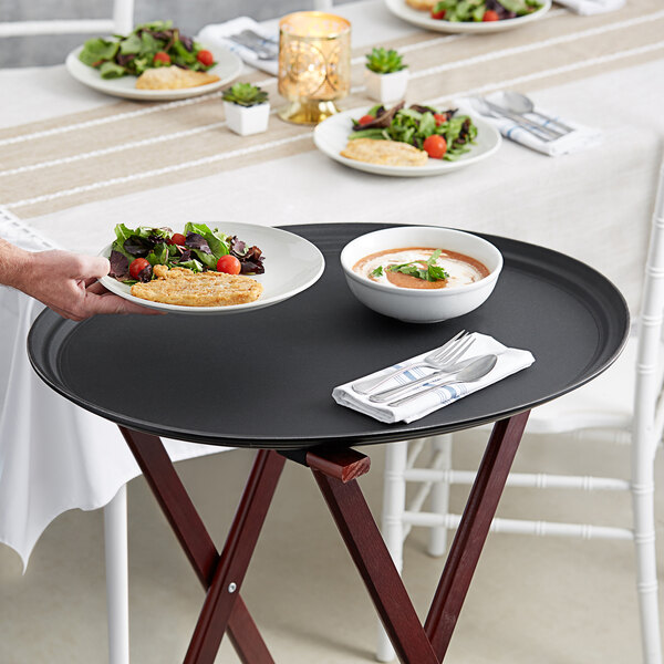 A person holding a Choice black oval non-skid serving tray with a bowl of soup and a plate of food on a table.