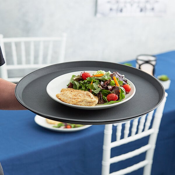 A person holding a Choice black non-skid serving tray with a plate of salad and chicken.