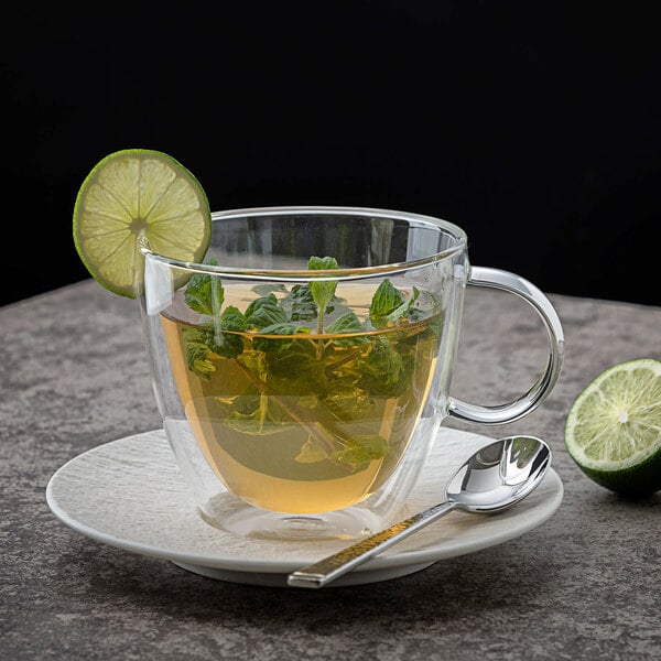 A Villeroy & Boch Artesano Barista double wall glass cup with tea and a lime wedge on the saucer with a spoon.