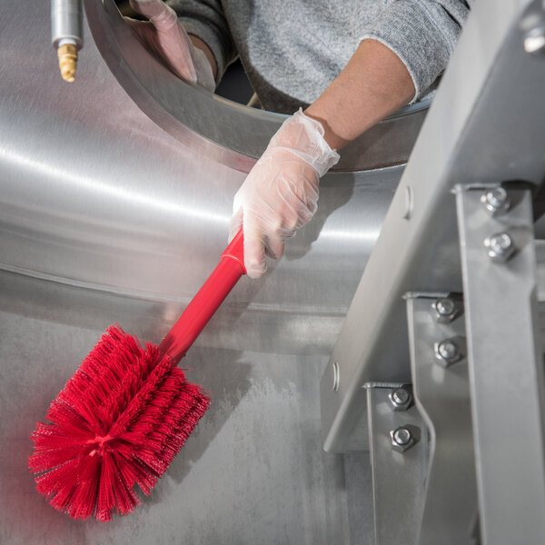 A person in gloves cleaning a metal object with a red Carlisle Sparta cleaning brush.