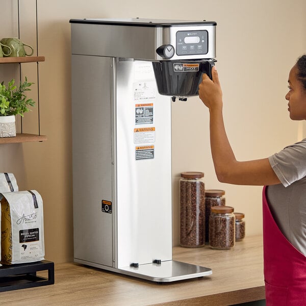 A woman using a Bunn BrewWISE stainless steel coffee maker on a counter in a coffee shop.
