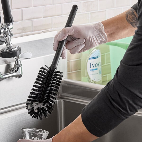 A person in rubber gloves using a Carlisle black bottle cleaning brush over a sink.