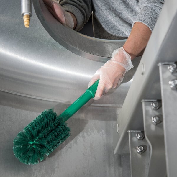 A hand holding a green Carlisle Sparta multi-purpose cleaning brush while cleaning a circular object.