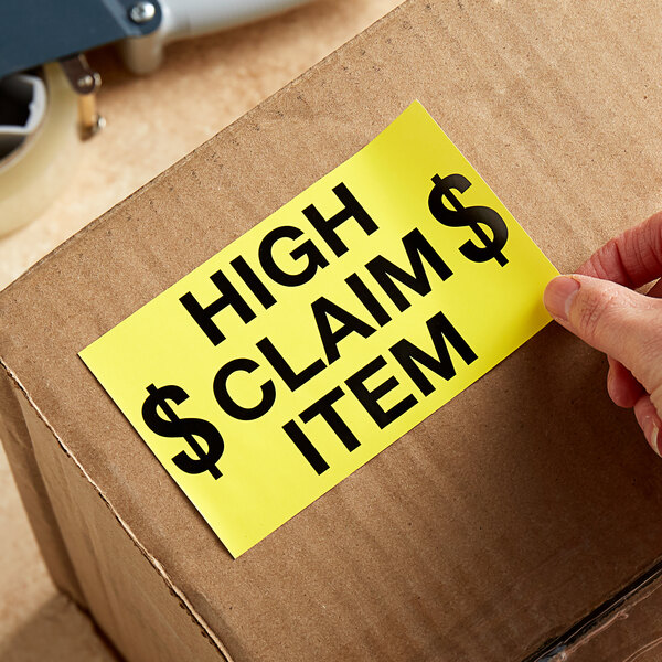 A hand using a Lavex High Claim Item label to mark a cardboard box.