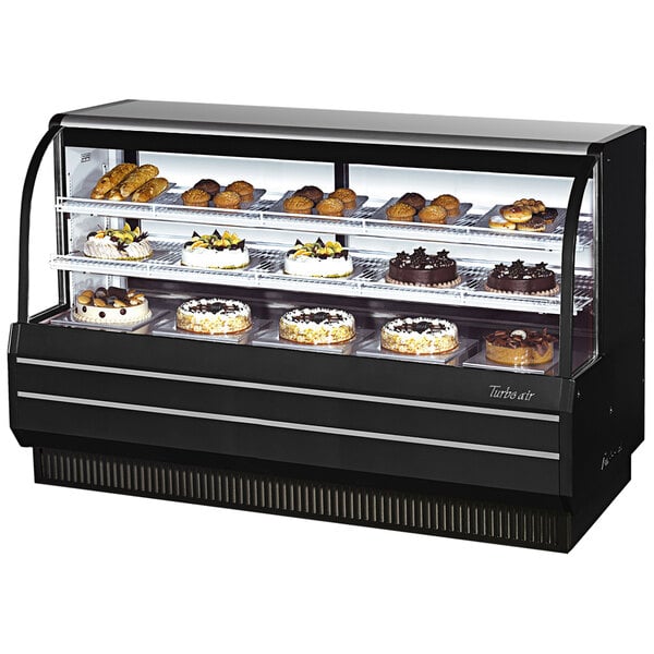 A Turbo Air black curved glass refrigerated bakery display case on a counter with cakes and pastries.