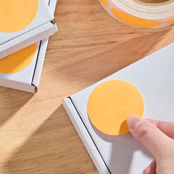 A roll of 2-inch fluorescent orange matte paper permanent round inventory labels being applied to white boxes.