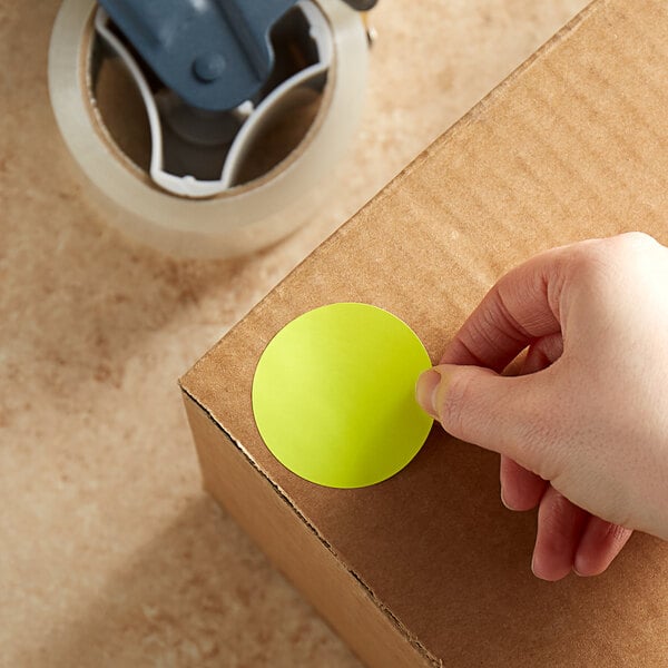 A hand using a Lavex round yellow sticker to label a cardboard box.