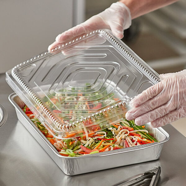 A person holding a ChoiceHD silver foil entree pan with a clear plastic lid over a tray of food.