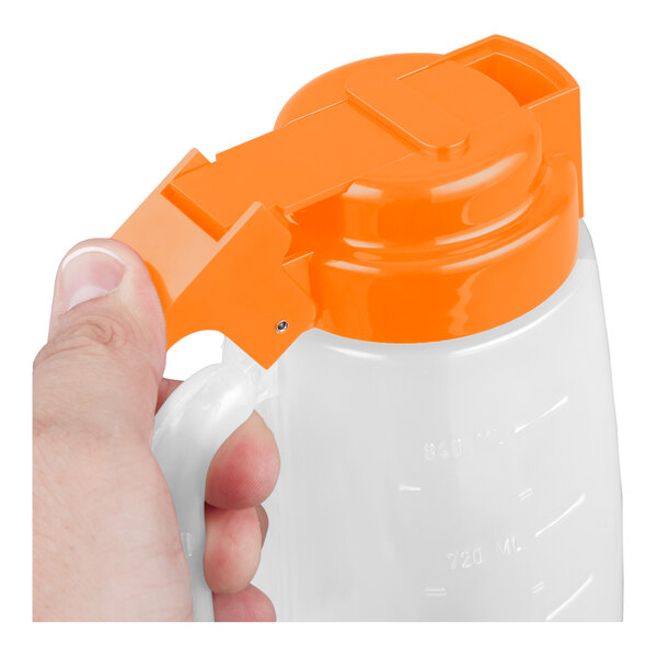 A clear plastic dispenser jar with an orange lid and handle, being held by a hand.