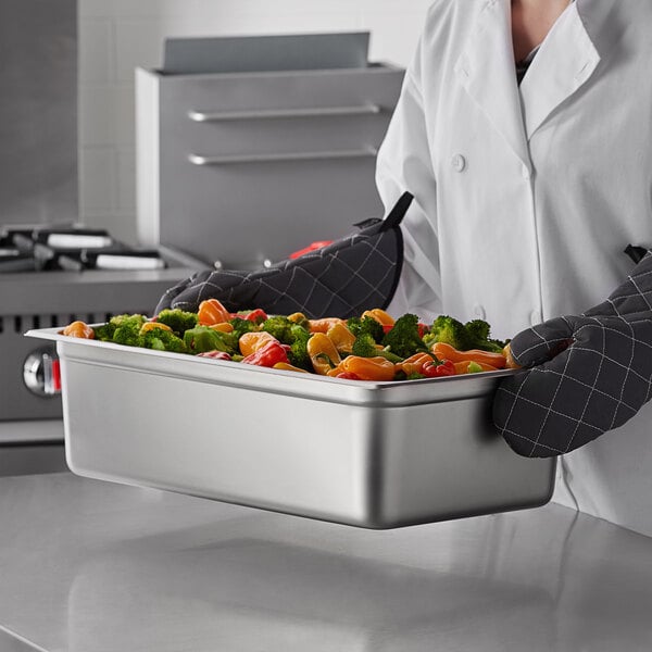 A person in a chef's uniform holding a Carlisle stainless steel steam table pan full of vegetables.