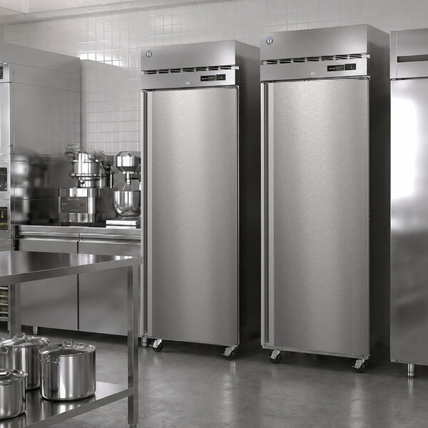 Two tall, stainless steel, single-door commercial freezers are positioned side by side in a professional kitchen setting with other stainless steel appliances and equipment visible.