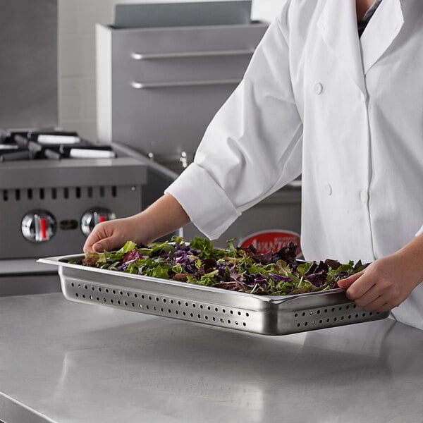 A woman in a white coat holding a Carlisle stainless steel perforated steam table pan full of salad.