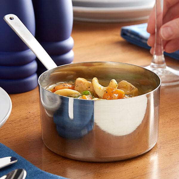 A person using a Vollrath mini stainless steel sauce pan to serve soup on a table.